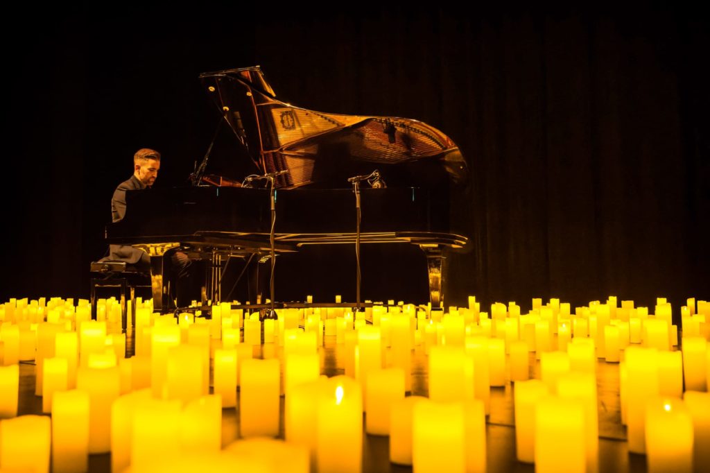 Un homme joue un concert de piano Candlelight au milieu de l'image avec un fond noir et des centaines de bougies à ses pieds.