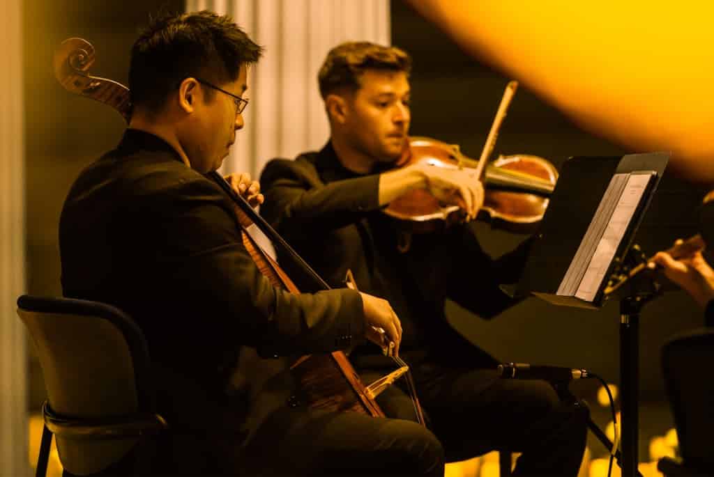 Deux hommes joue du violon et du violoncelle lors d'un concert Candlelight sous la lumière des bougies face à des pupitres et des partitions.