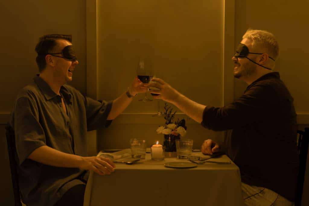 Photo de deux hommes trinquant leurs verres de vins au tour d'une table partageant un dîner à l'aveugle Dining in the Dark